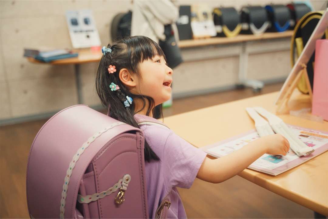 ライラック色のランドセルを背負った女の子が、色見本に手をのばして笑っている